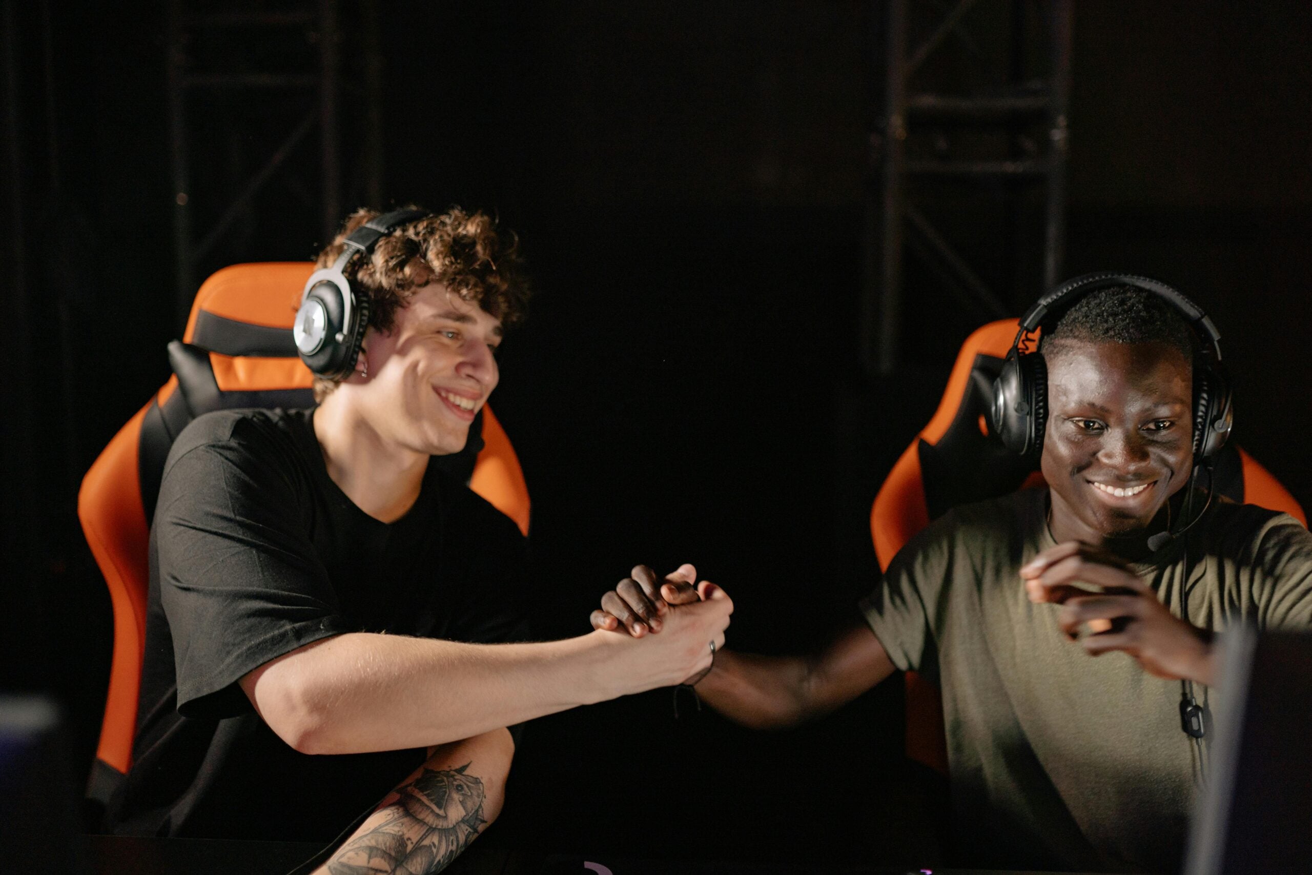 Two gamers wearing headsets sit in orange gaming chairs, smiling and shaking hands in a darkened room after a match.
