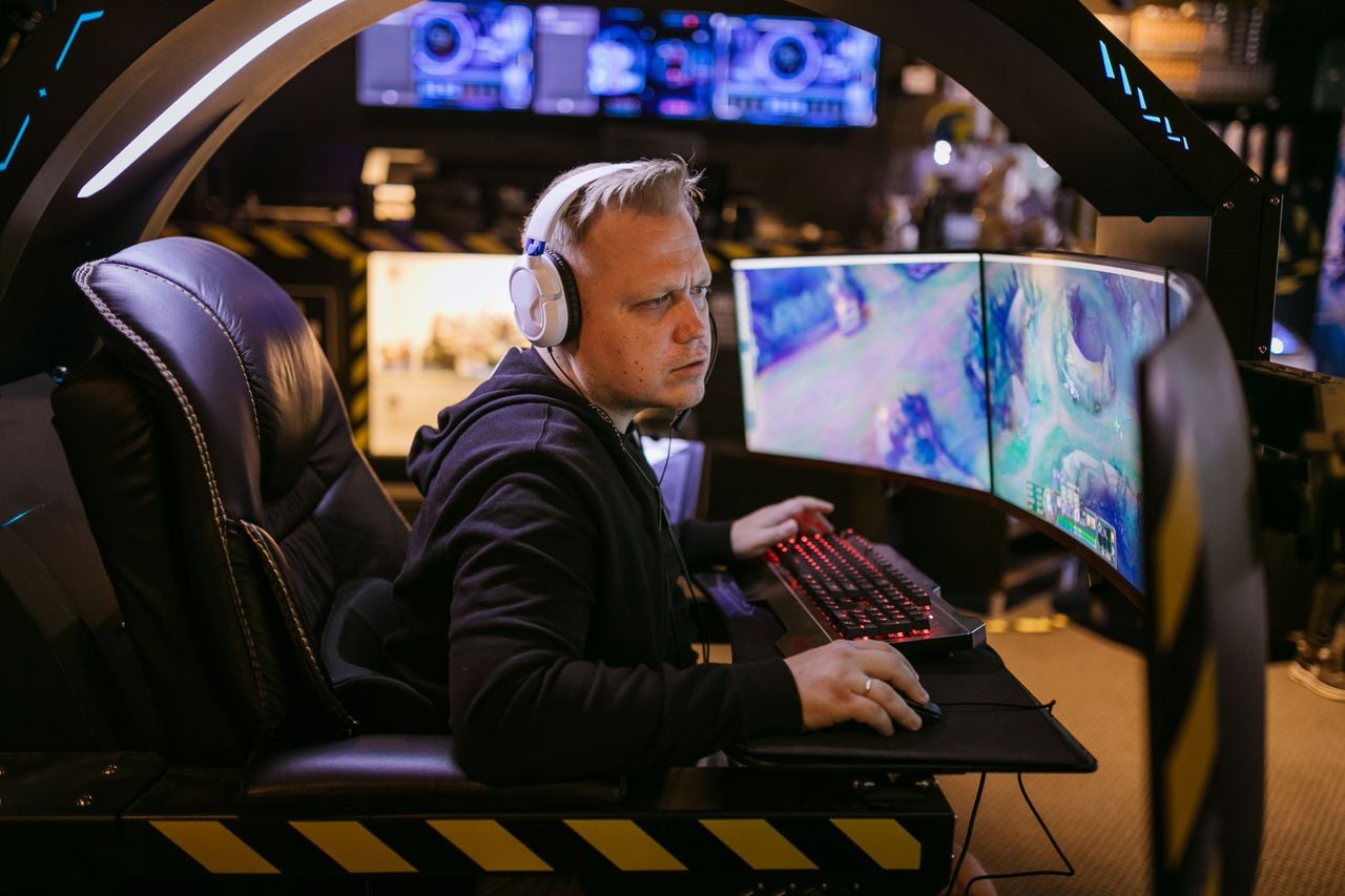 A gamer wearing a headset plays a computer game at a multi-monitor gaming setup, with colorful game screens and RGB-lit peripherals in the background.